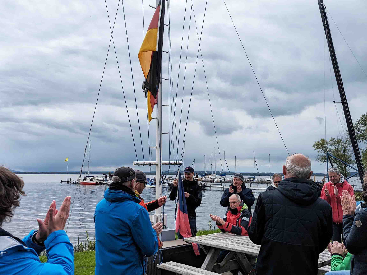 Es ist der Flaggenmast zu sehen und wie zwei Mitglieder die Klubfahne und eine Deutschlandfahne befestigen, um sie hoch zu ziehen. Um den Flaggenmast herum haben sich weitere Mitglieder versammelt, die alle klatschen.