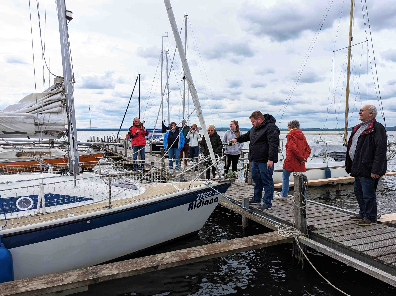 Eine Person steht auf einem Steg vor einem Schiff und schüttet den Inhalt einer Sektflasche auf das Schiff. Auf dem Steg stehen weitere Personen, die die Szene gespannt beobachten.